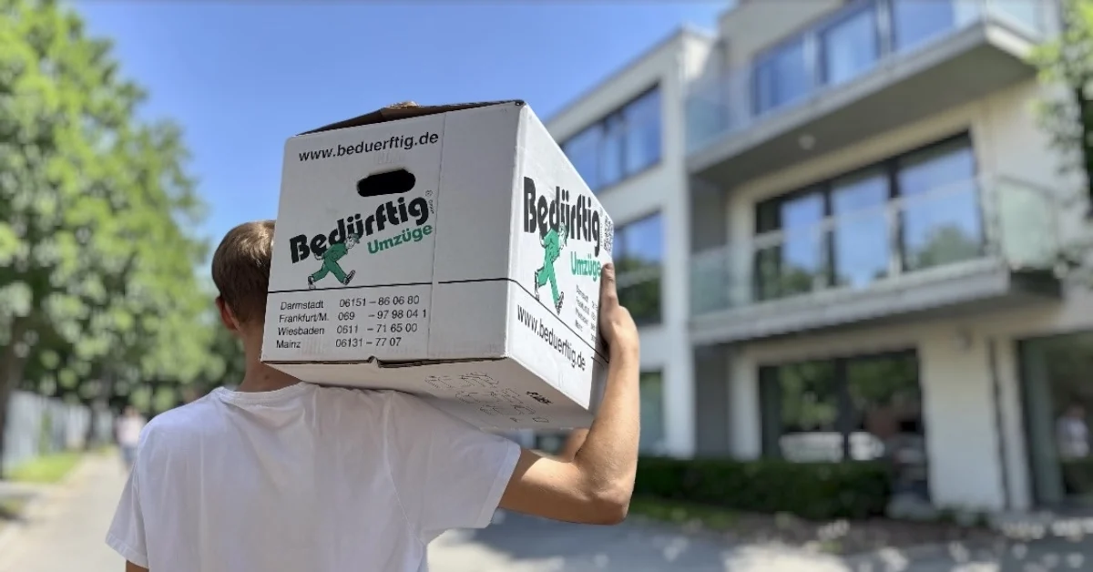 Mitarbeiter trägt einen Umzugskarton zu einer Wohnanlage